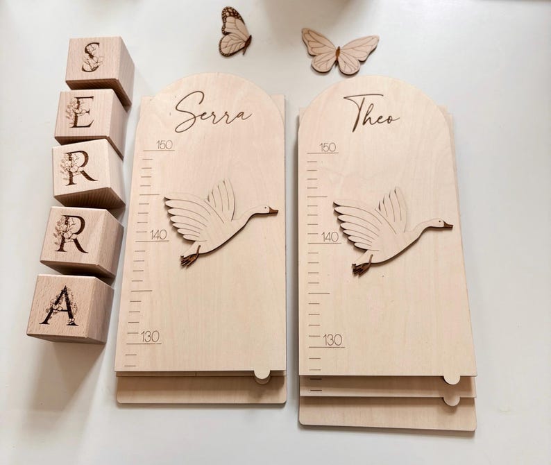K&ouml;nnte beinhalten: Wachstumstafel aus Holz mit einem fliegenden G&auml;nsemotiv, misst bis zu 150 cm. Die Tafel ist mit den Namen "Serra" und "Theo" personalisiert. Holzbl&ouml;cke buchstabieren den Namen "SERRA". Dekorative Schmetterlinge sind ebenfalls vorhanden.