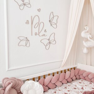 May include: A nursery scene featuring a white crib with a pink braided bumper and floral bedding. Wooden butterfly wall decor and the name "Pola" are above the crib. A canopy with swan decorations hangs above the crib, creating a soft, inviting space.