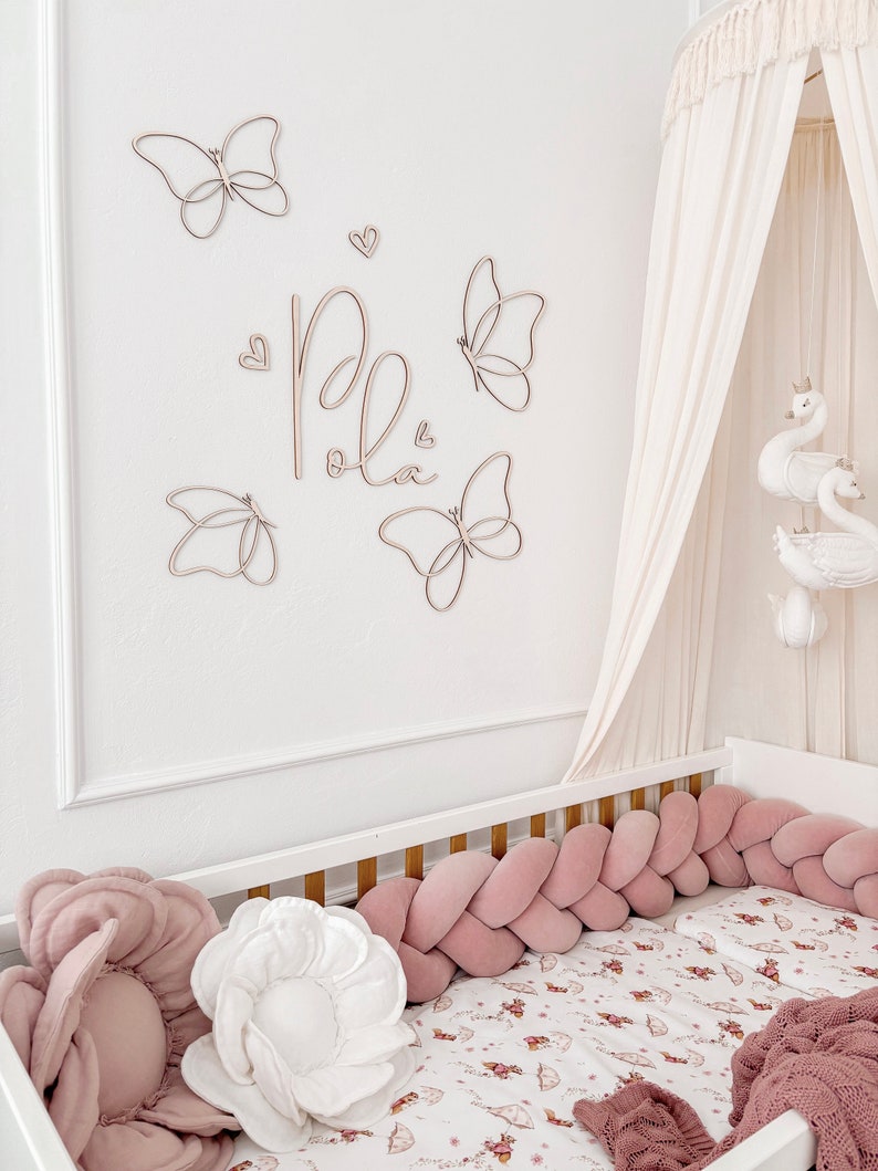 May include: A nursery scene featuring a white crib with a pink braided bumper and floral bedding. Wooden butterfly wall decor and the name "Pola" are above the crib. A canopy with swan decorations hangs above the crib, creating a soft, inviting space.