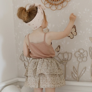 May include: A young child wearing a pink tank top, floral print tiered skirt, and a matching headband. The child is reaching towards wooden butterfly and flower cutouts on a floral wallpaper background. A rattan mirror is visible.