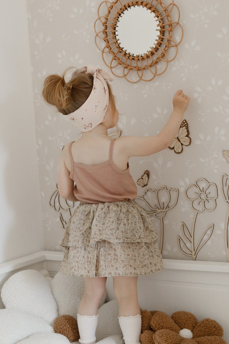 May include: A young child wearing a pink tank top, floral print tiered skirt, and a matching headband. The child is reaching towards wooden butterfly and flower cutouts on a floral wallpaper background. A rattan mirror is visible.