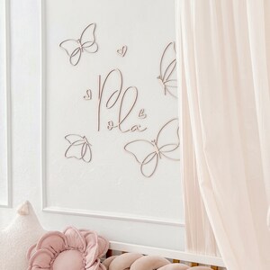 May include: A wall decoration featuring wooden butterflies and the name "Pola" in cursive script. The butterflies and hearts are arranged on a white wall, with a soft pink flower pillow and a braided bumper visible.