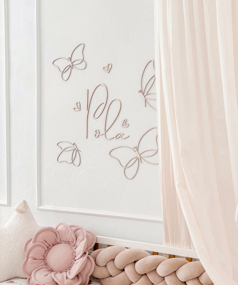 May include: A wall decoration featuring wooden butterflies and the name "Pola" in cursive script. The butterflies and hearts are arranged on a white wall, with a soft pink flower pillow and a braided bumper visible.