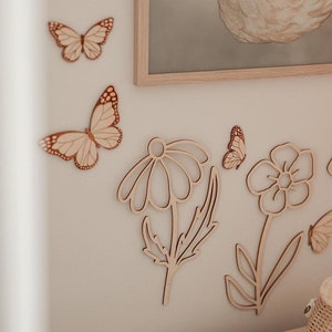 May include: Wooden wall decor featuring butterflies and flowers. The butterflies have detailed wing patterns, and the flowers are stylized with open petals and stems. The pieces are made of light-colored wood and are mounted on a neutral-toned wall.