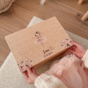 Op de afbeelding: Een houten herinneringsdoos met een ballerina-illustratie en bloemmotieven. De doos is voorzien van de naam "Jena" en de datum "13.11.2019". De doos wordt vastgehouden door een persoon die een crèmekleurige trui en een roze tutu draagt.