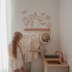 May include: A child's bedroom scene with a play kitchen, dresses, and decorative wall art. The wall features wooden flowers and butterflies. Two dresses hang on a rack. A woven basket with bear ears is on the wall. The play kitchen has a copper faucet.