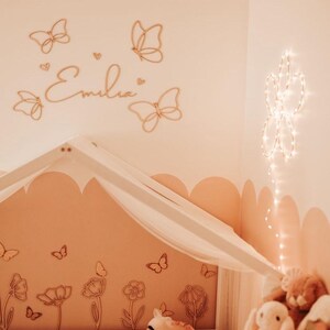 May include: A nursery with a soft peach color scheme. Wooden butterfly wall decor and the name "Emilia" are above a canopy bed. A floral mural and plush toys add to the room's whimsical feel. String lights in a flower shape are on the wall.