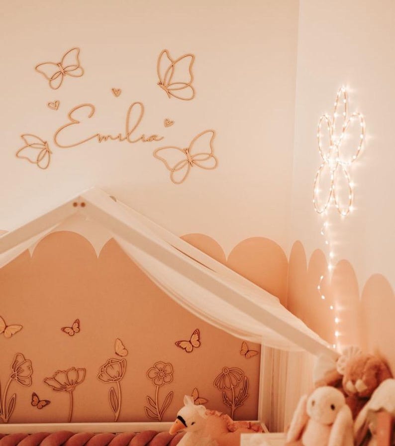 May include: A nursery with a soft peach color scheme. Wooden butterfly wall decor and the name "Emilia" are above a canopy bed. A floral mural and plush toys add to the room's whimsical feel. String lights in a flower shape are on the wall.