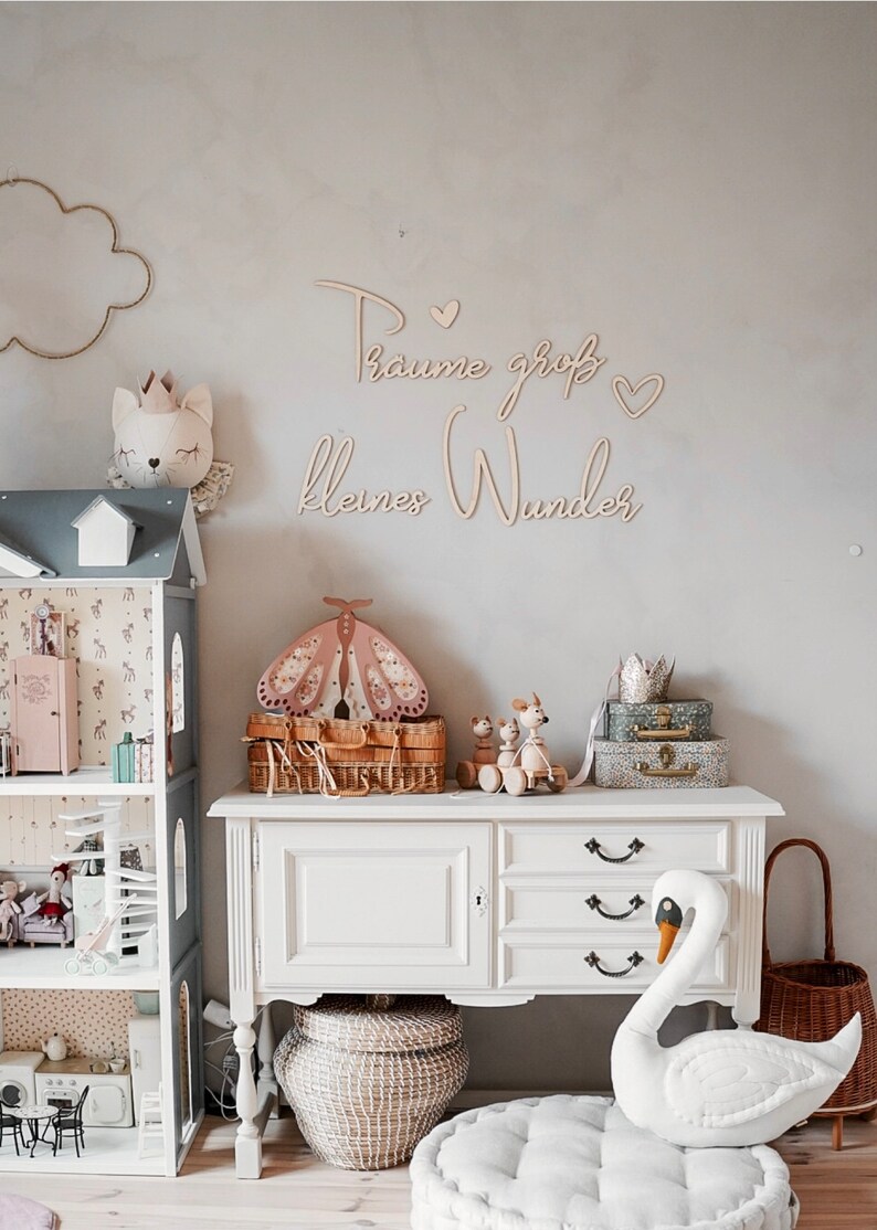 May include: A white dresser with three drawers and a white swan plush toy sitting on a gray cushion. The dresser is decorated with a wooden sign that reads "Traume gro&szlig; kleines Wunder" and a wooden butterfly.