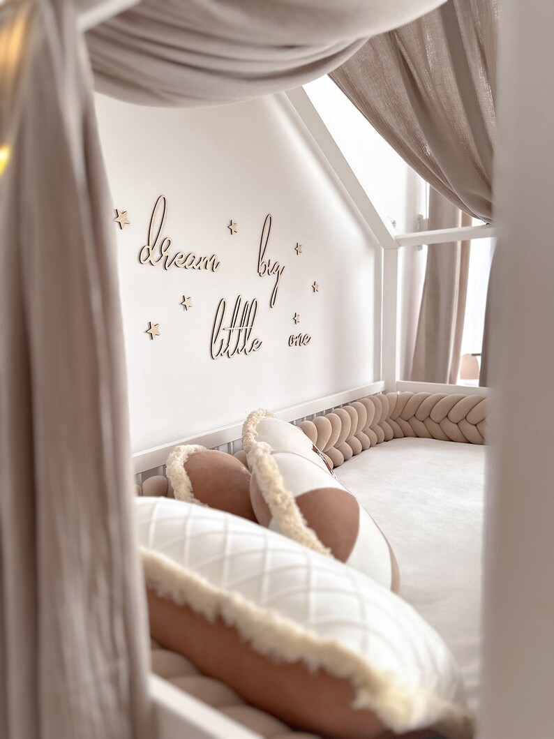 K&ouml;nnte beinhalten: Ein wei&szlig;es Himmelbett mit einem geflochtenen, beigen Sto&szlig;f&auml;nger und einem gesteppten Kissen in Wei&szlig; und Beige. Die Wand hinter dem Bett hat die Worte "dream big little one" in Kursivschrift mit Holzsternen geschrieben.