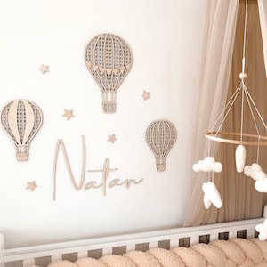 Puede incluir: Una cuna blanca con un parachoques trenzado beige y un dosel blanco. La pared detr&aacute;s de la cuna est&aacute; decorada con globos aerost&aacute;ticos de madera, estrellas y el nombre "Natan".