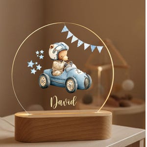 May include: A decorative night light featuring a teddy bear in a blue race car, wearing a helmet. The design includes blue stars and a banner, with the name "David" in script. The light sits on a wooden base.