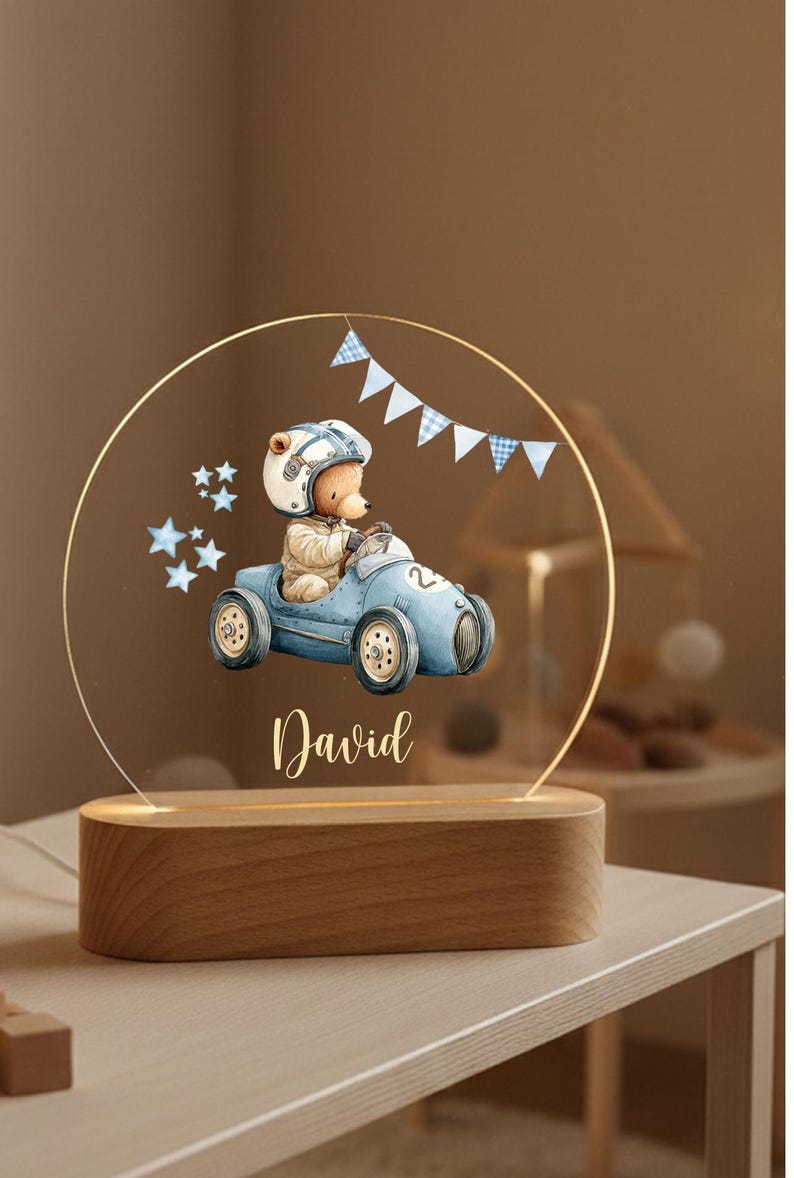 May include: A decorative night light featuring a teddy bear in a blue race car, wearing a helmet. The design includes blue stars and a banner, with the name "David" in script. The light sits on a wooden base.