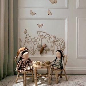 May include: Two dolls sit at a small table set for tea. The dolls are dressed in patterned dresses and hats. The table and chairs are made of light-colored wood. Wooden butterflies and flower outlines decorate the wall behind them.