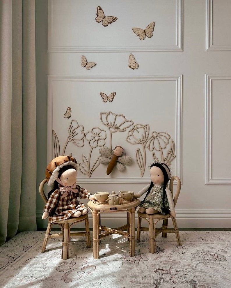 May include: Two dolls sit at a small table set for tea. The dolls are dressed in patterned dresses and hats. The table and chairs are made of light-colored wood. Wooden butterflies and flower outlines decorate the wall behind them.