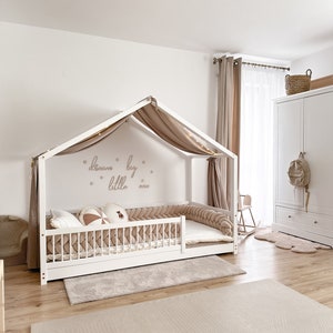 K&ouml;nnte beinhalten: Ein wei&szlig;es Holz-Baldachinbett mit einem beigefarbenen Baldachin und einem beigefarbenen geflochtenen Sto&szlig;f&auml;nger. Das Bett hat eine wei&szlig;e Matratze und einen beigefarbenen Teppich darunter. Die Wand hinter dem Bett hat einen beigefarbenen Aufkleber mit der Aufschrift "dream big little one".