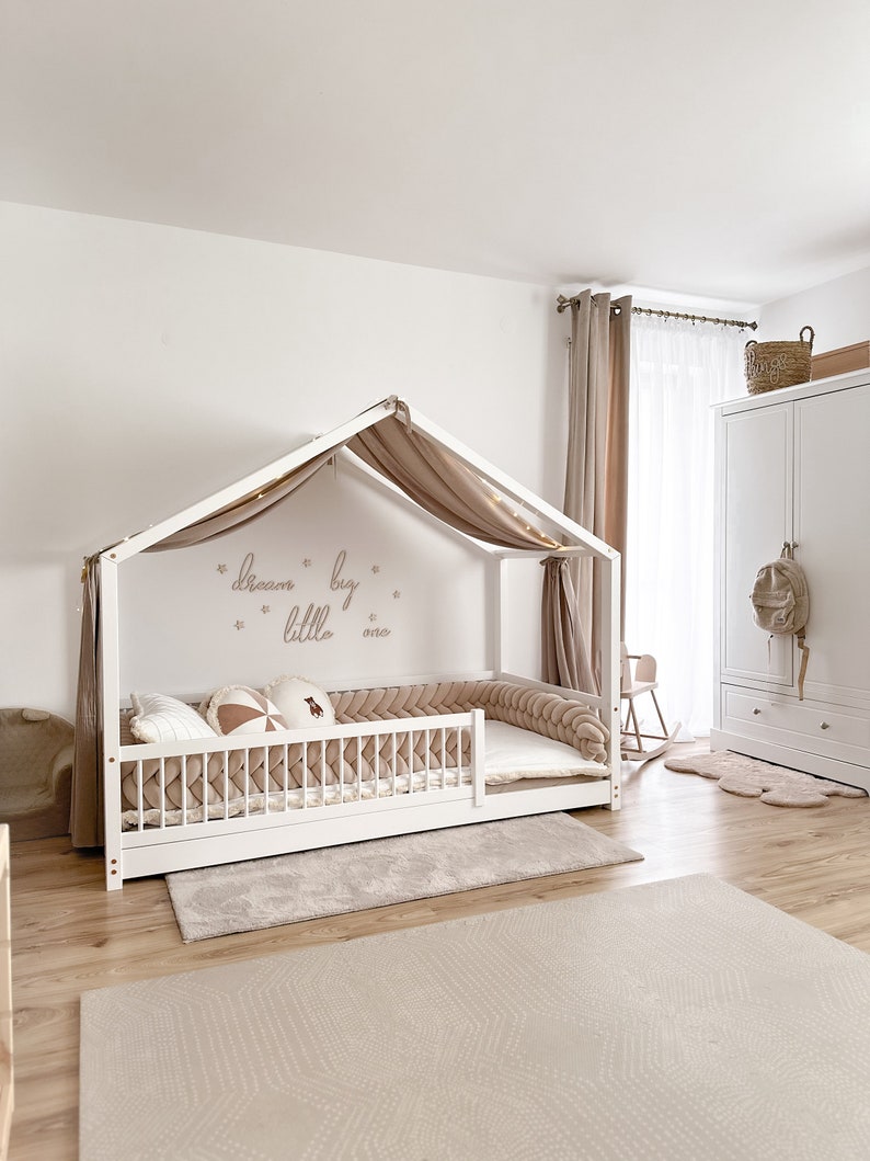 K&ouml;nnte beinhalten: Ein wei&szlig;es Holz-Baldachinbett mit einem beigefarbenen Baldachin und einem beigefarbenen geflochtenen Sto&szlig;f&auml;nger. Das Bett hat eine wei&szlig;e Matratze und einen beigefarbenen Teppich darunter. Die Wand hinter dem Bett hat einen beigefarbenen Aufkleber mit der Aufschrift "dream big little one".