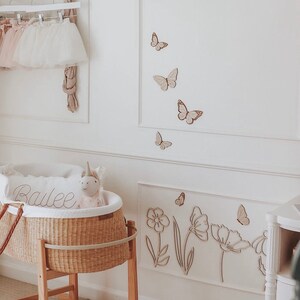 May include: A nursery scene with a wicker bassinet on a wooden stand, a pillow with the name "Bailee", and a stuffed unicorn. The white walls are decorated with wooden butterflies and floral cutouts. A rack holds tutus.