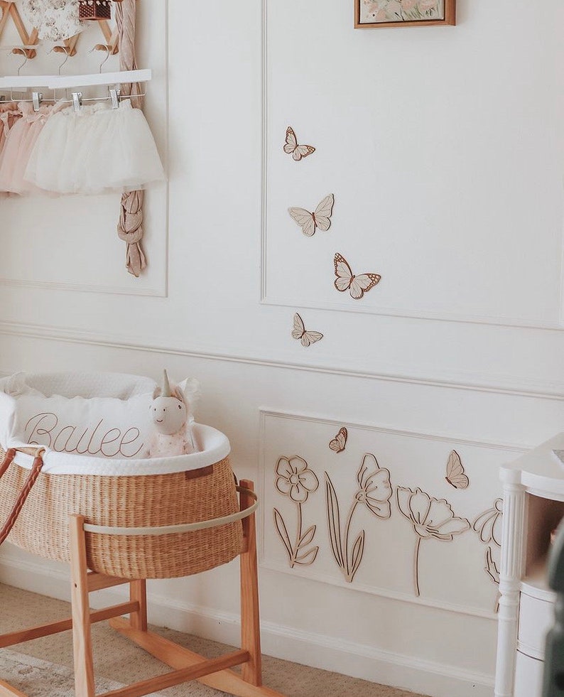 May include: A nursery scene with a wicker bassinet on a wooden stand, a pillow with the name "Bailee", and a stuffed unicorn. The white walls are decorated with wooden butterflies and floral cutouts. A rack holds tutus.