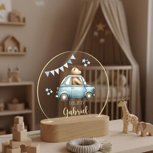 May include: A decorative night light featuring a blue car with a teddy bear driver, a banner, and stars. The name "Gabriel" and the date "1.06.2022" are displayed below the car. The light sits on a wooden base.