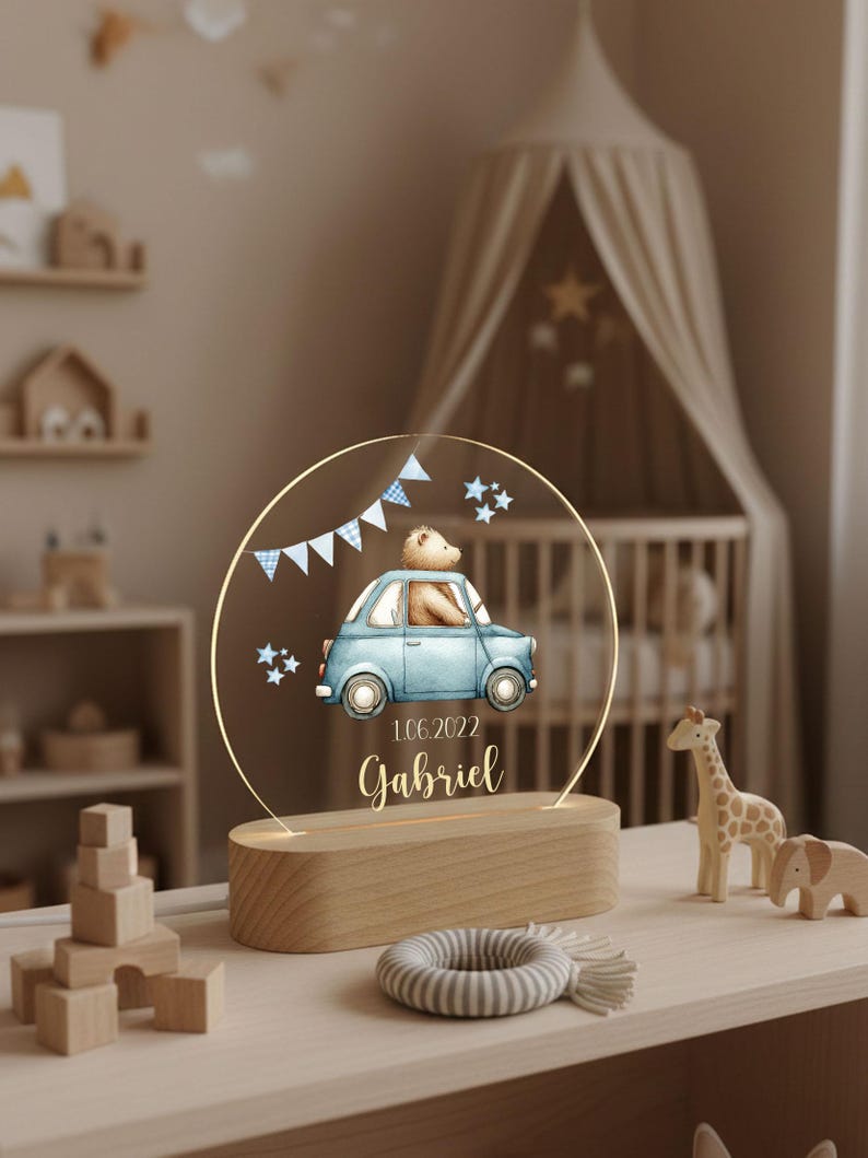May include: A decorative night light featuring a blue car with a teddy bear driver, a banner, and stars. The name "Gabriel" and the date "1.06.2022" are displayed below the car. The light sits on a wooden base.