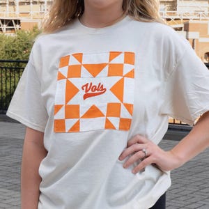 Pode incluir: Uma t-shirt de manga curta na cor creme com um grande emblema quadrado no peito. O emblema apresenta um padrão geométrico tipo colcha em laranja brilhante e branco, com a palavra "Vols" bordada em letra cursiva vermelha no centro.