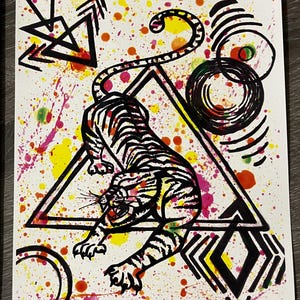 May include: A vibrant artwork featuring a black and white tiger within a black triangle, set against a white background with colorful paint splatters. The tiger has black stripes and is in a crouching pose. Geometric shapes and abstract designs surround the tiger.