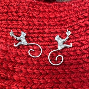 Puede incluir: Dos porta-bolas de lana de mono plateados sobre un fondo de punto rojo. Los monos sostienen una bola de lana en sus manos.