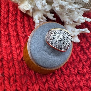 May include: A silver ring with an ornate, swirling design and a gold band. The ring is displayed on a gray ring holder, set against a red knitted background and white coral.