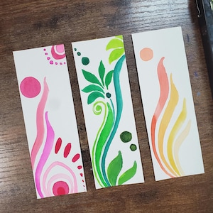 Peut inclure: Trois marque-pages aquarelle avec des motifs abstraits. L'un présente des tourbillons roses et rouges avec un cercle rose. Un autre a des feuilles vertes et une tige turquoise. Le dernier a des formes de flammes orange et jaunes.