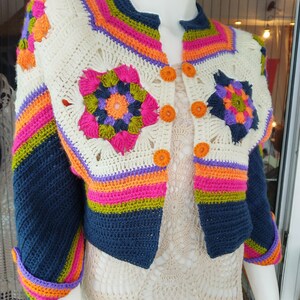May include: A crocheted cardigan with a colorful striped pattern and floral motifs. The cardigan has a button closure and is made with a variety of colors, including pink, orange, green, blue, and white.