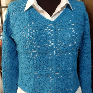 Könnte beinhalten: Ein türkisfarbener Häkelpullover mit V-Ausschnitt und langen Ärmeln. Der Pullover ist mit einem quadratischen Stichmuster gearbeitet und hat eine lockere Passform.