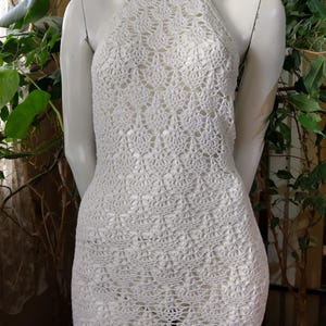 Puede incluir: Vestido blanco de crochet con cuello halter, exhibido en un maniquí. El vestido presenta un patrón calado detallado y una silueta ajustada. El vestido es de color blanco liso y parece hecho a mano.