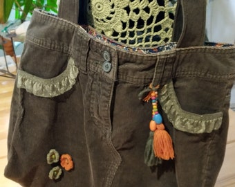 Pode incluir: Uma bolsa tote de veludo cotelê marrom com uma borda de crochê e um tassel de contas coloridas. A bolsa tem um bolso grande na frente e é decorada com pequenas flores de tecido.