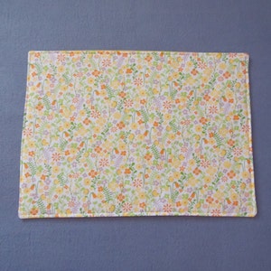 Könnte beinhalten: Ein weißer Platzdeckchen mit einem Blumenmuster mit kleinen gelben, orangefarbenen und lila Blüten auf grünen Stielen. Das Platzdeckchen hat einen hellorangen Rand.