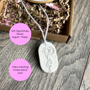 May include: An oval white clay gift tag featuring an embossed poppy flower design. The tag is suspended by a light gray string and is positioned in front of a brown cardboard box filled with crinkled gold and pink paper filler material.