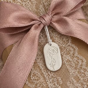 A small oval shaped gift tag handmade from air dry clay. The tag is stamped with a poppy flower representing the birth flower for August .  the gift tag can be purchased with a cotton drawstring bag . other items are available to match.