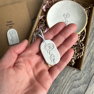May include: A white clay gift set featuring a round trinket dish with a gold rim, an oval pendant with a floral imprint, and a rectangular card with the text Happy Birthday Reb, all packaged in a brown cardboard box with paper filler.
