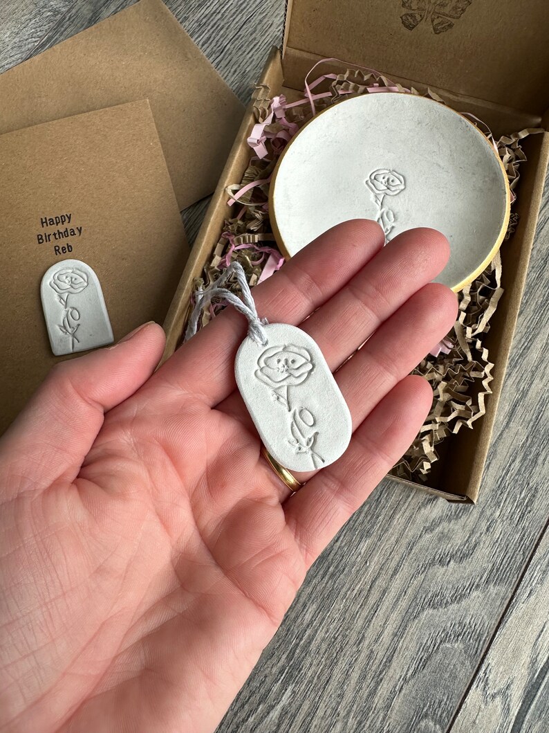 May include: A white clay gift set featuring a round trinket dish with a gold rim, an oval pendant with a floral imprint, and a rectangular card with the text Happy Birthday Reb, all packaged in a brown cardboard box with paper filler.