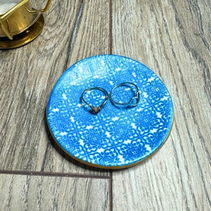 May include: A small, round blue and white patterned ring dish with two rings. One ring is gold with a small stone, and the other is silver with a heart-shaped design. The dish is on a wooden surface.