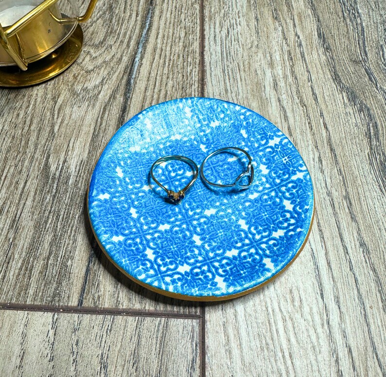 May include: A small, round blue and white patterned ring dish with two rings. One ring is gold with a small stone, and the other is silver with a heart-shaped design. The dish is on a wooden surface.