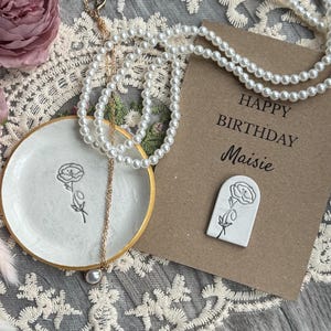 May include: A white ceramic trinket dish with a gold rim and a black floral line drawing, a gold chain necklace with a single pearl pendant, and a brown card with the text Happy Birthday Maisie and a white arch-shaped floral pin.