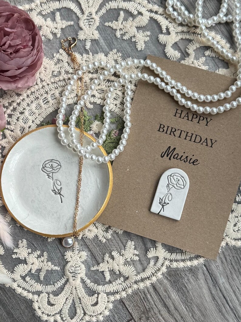 May include: A white ceramic trinket dish with a gold rim and a black floral line drawing, a gold chain necklace with a single pearl pendant, and a brown card with the text Happy Birthday Maisie and a white arch-shaped floral pin.