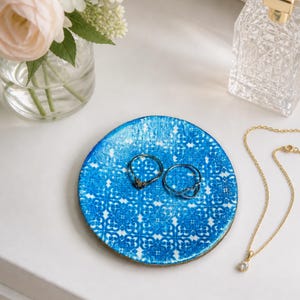 May include: A round, blue and white patterned trinket dish holds two rings. A gold necklace with a small pendant and a perfume bottle are nearby. A vase of flowers sits in the background.