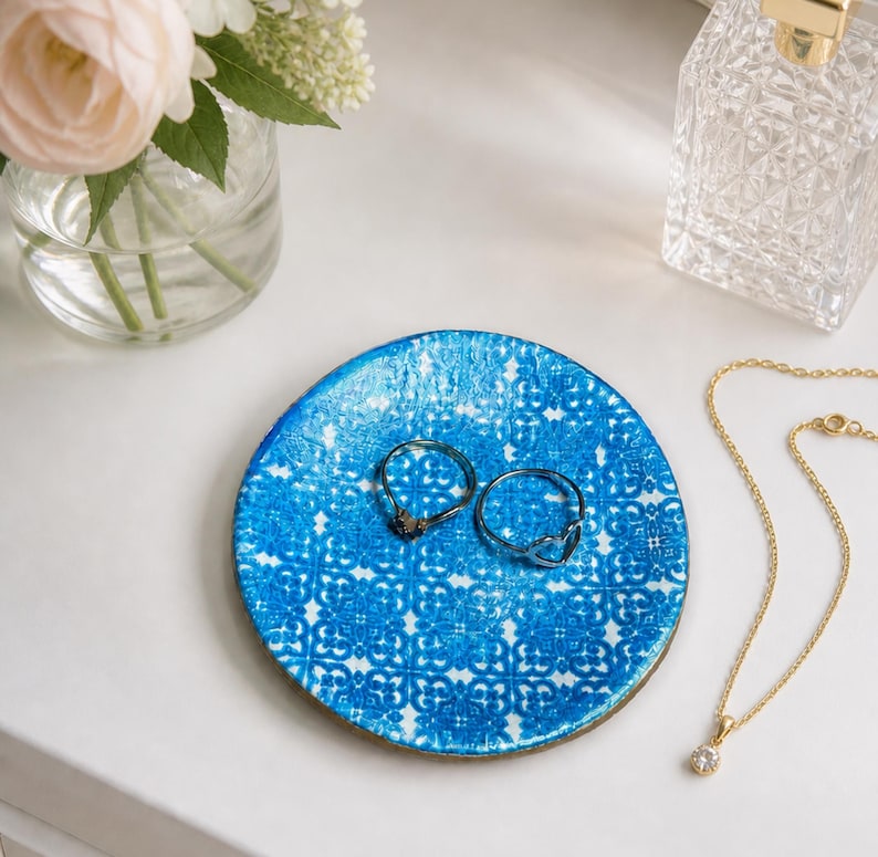 May include: A round, blue and white patterned trinket dish holds two rings. A gold necklace with a small pendant and a perfume bottle are nearby. A vase of flowers sits in the background.