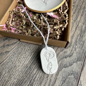 May include: A white clay trinket dish with a gold-painted rim and a matching oval clay tag, both featuring an embossed rose design. The tag is attached to a white string. These items are nestled in a brown cardboard box with shredded paper.