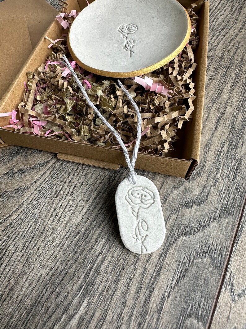 May include: A white clay trinket dish with a gold-painted rim and a matching oval clay tag, both featuring an embossed rose design. The tag is attached to a white string. These items are nestled in a brown cardboard box with shredded paper.