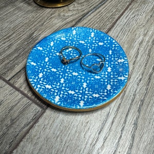May include: A small, round blue and white patterned ceramic dish with a gold rim, holding two silver rings. One ring has a heart shape, and the other has a small stone. A gold candle holder is in the background.