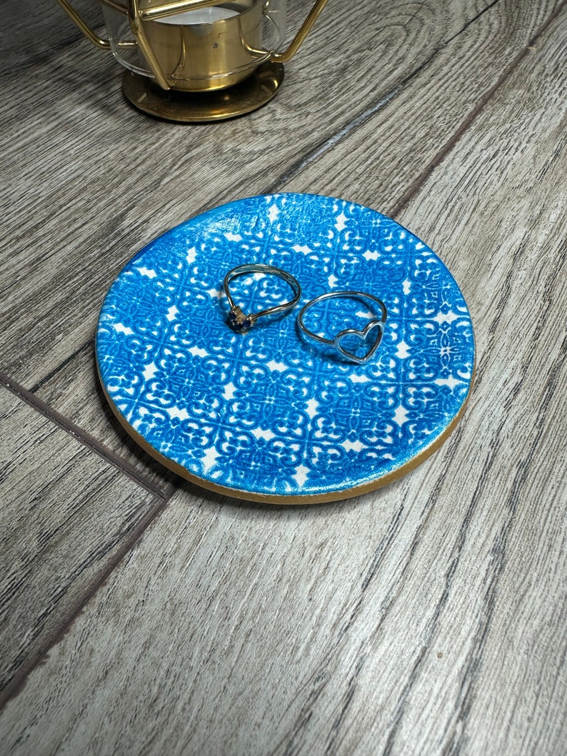 May include: A small, round blue and white patterned ceramic dish with a gold rim, holding two silver rings. One ring has a heart shape, and the other has a small stone. A gold candle holder is in the background.
