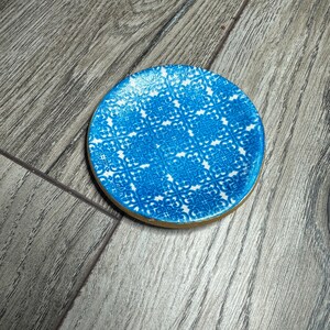 May include: A round coaster with a blue and white patterned design. The coaster has a gold-colored edge. The pattern is a repeating geometric design. The coaster is sitting on a wooden surface.